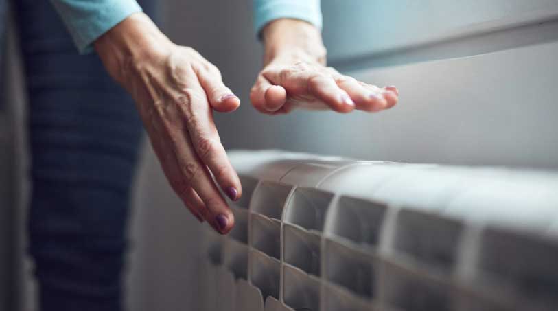 Woman's hands hovering over a white radiator to feel warmth, suggesting heating system usage during cold weather.